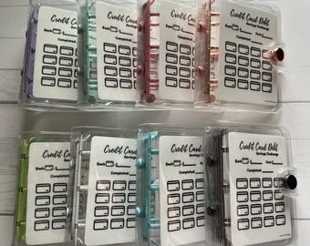 May include: Six clear plastic binders with colorful rings and white inserts. Each insert has a title that reads "Credit Card Savings Challenge" or "Credit Card Debt Savings Challenge". The inserts have a grid of squares with lines to check off.