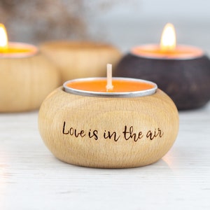 Könnte beinhalten: Drei Holz-Kerzenhalter mit Teelichtern. Ein Halter ist braun und die anderen beiden sind aus Naturholz. Der Halter in der Mitte hat den Text "Love is in the air" eingraviert.