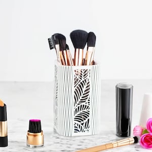 May include: White makeup brush holder with a leaf design. The holder is filled with various makeup brushes with black and gold handles.