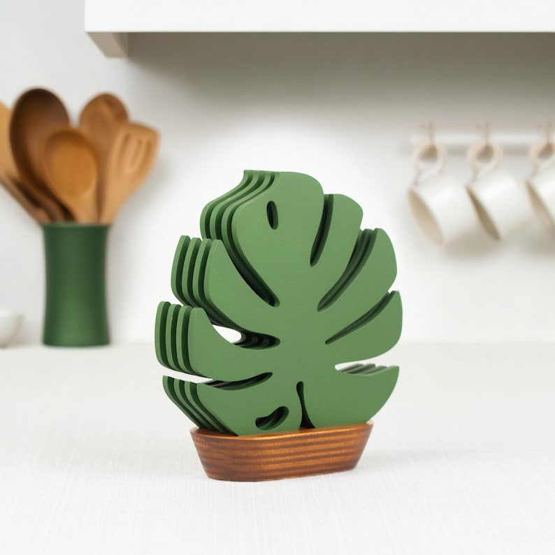 May include: Set of six green monstera leaf-shaped coasters with a wooden base. The coasters are stacked on top of each other and are arranged in a fan shape.