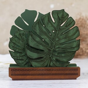 May include: Green wooden napkin holder with a carved design of tropical leaves. The holder has a brown base with a geometric pattern.