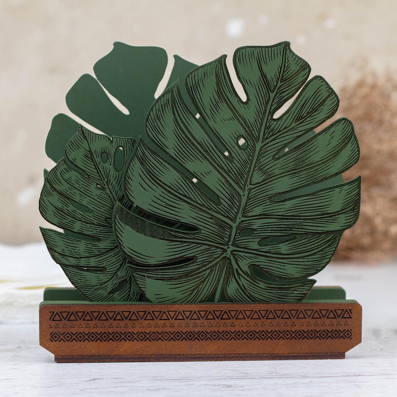 May include: Green wooden napkin holder with a carved design of tropical leaves. The holder has a brown base with a geometric pattern.