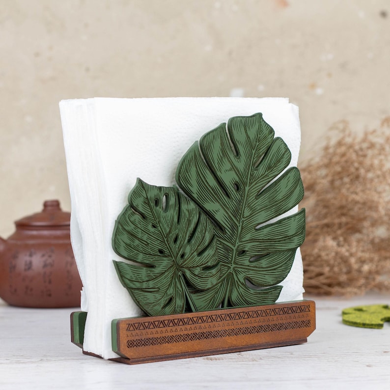 May include: Wooden napkin holder with a green monstera leaf design. The holder has a brown base with a geometric pattern.