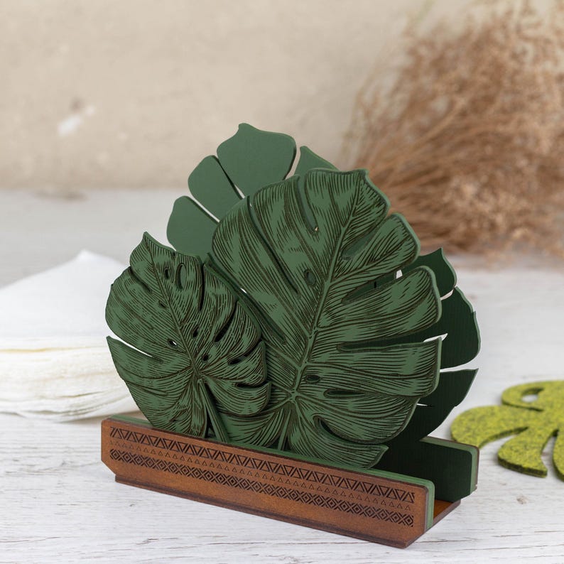 May include: Green wooden napkin holder shaped like tropical monstera leaves. The holder has a brown base with a geometric pattern.
