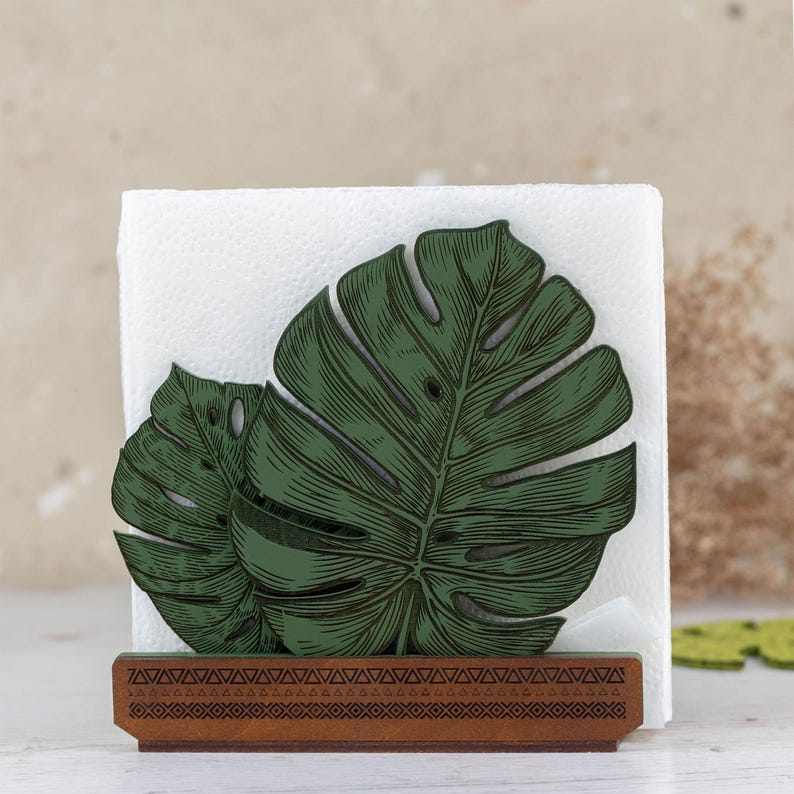 May include: Green wooden napkin holder shaped like a monstera leaf. The holder has a brown wooden base with a decorative pattern.