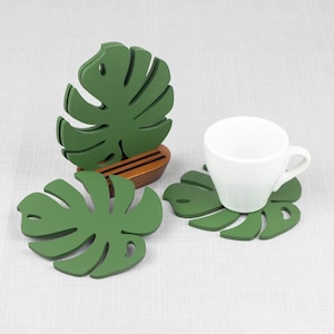 May include: Set of four green monstera leaf-shaped coasters with a white ceramic cup on one of the coasters. The coasters are made of wood and have a natural wood finish.