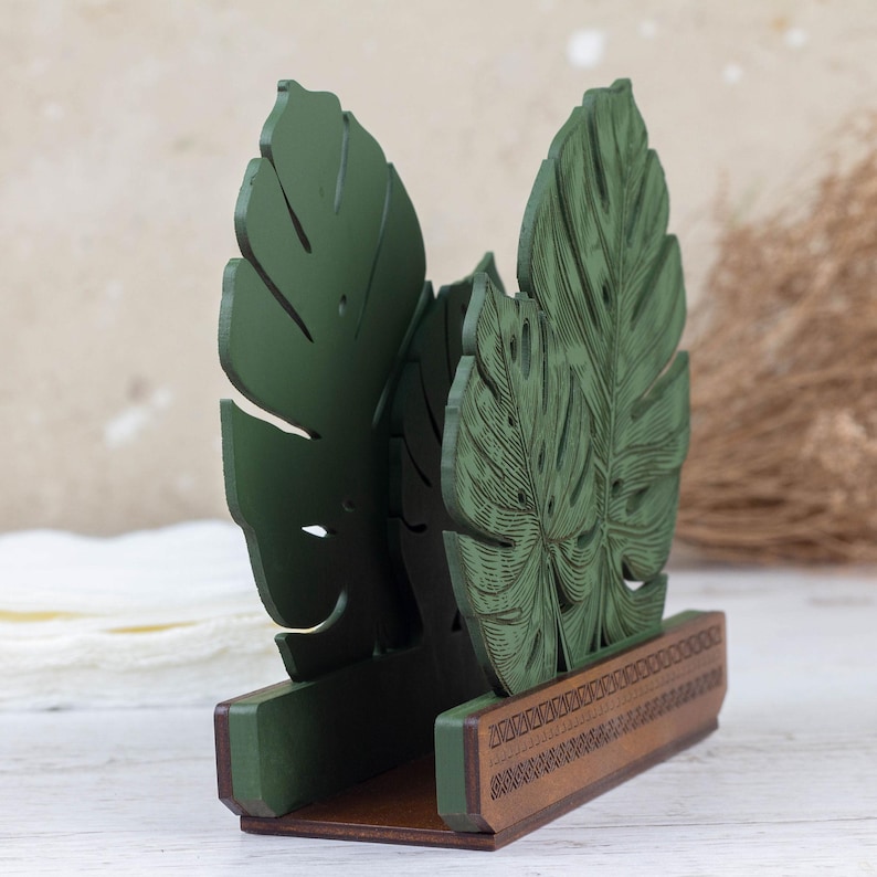 May include: Green wooden napkin holder shaped like two tropical leaves. The holder has a brown base with a decorative pattern.