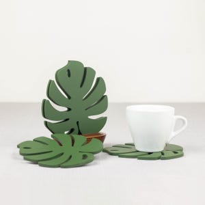 May include: Three green leaf-shaped coasters with a white ceramic cup on one of the coasters. The coasters are shaped like monstera leaves.