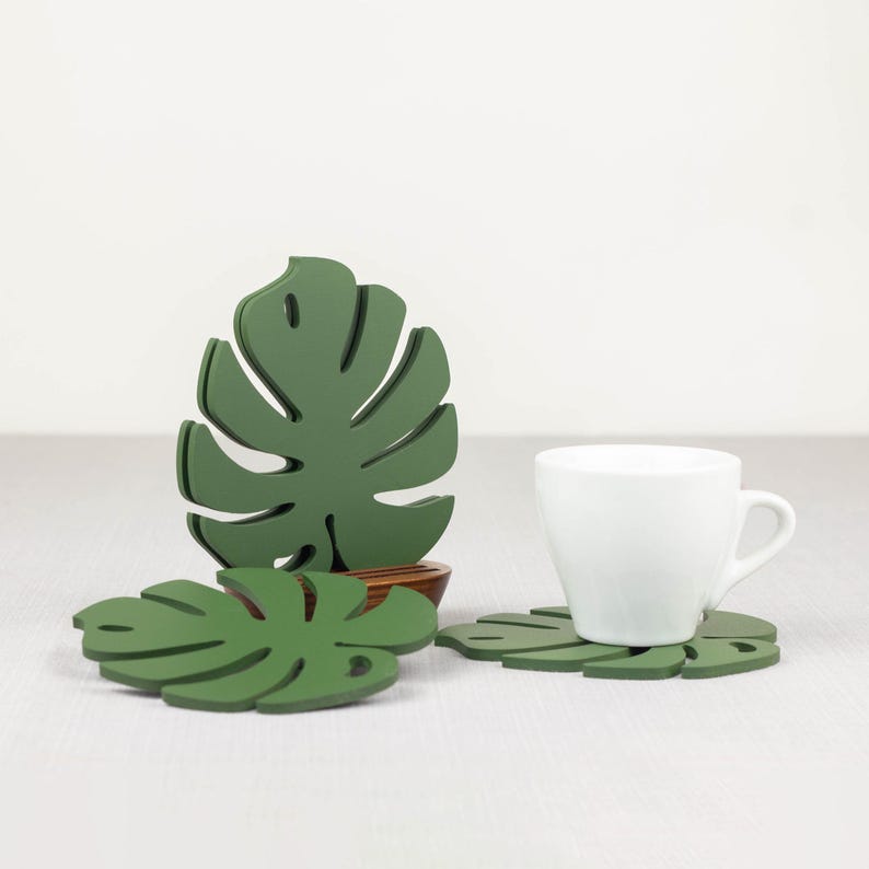 May include: Three green leaf-shaped coasters with a white ceramic cup on one of the coasters. The coasters are shaped like monstera leaves.