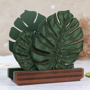 May include: Green wooden napkin holder shaped like two monstera leaves with a brown wooden base with a geometric pattern.