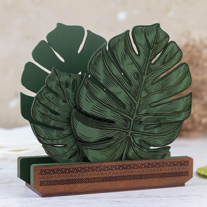 May include: Green wooden napkin holder shaped like two monstera leaves with a brown wooden base with a geometric pattern.