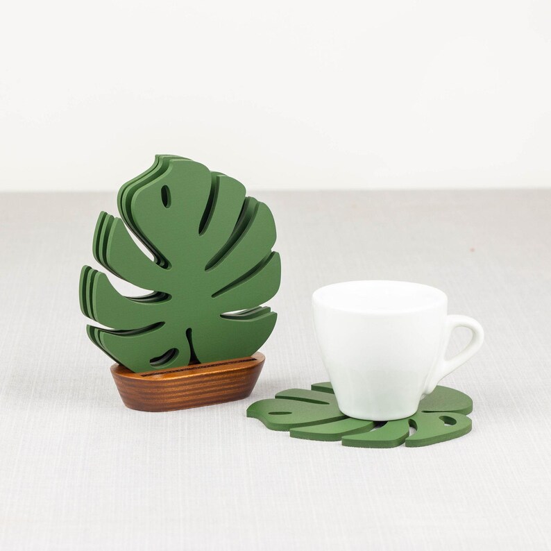 May include: Set of four green monstera leaf-shaped coasters with a wooden base. The coasters are stacked and the top coaster is sitting on a white cup.