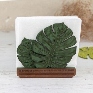 May include: A wooden napkin holder with a green monstera leaf design. The holder has a brown base with a geometric pattern.