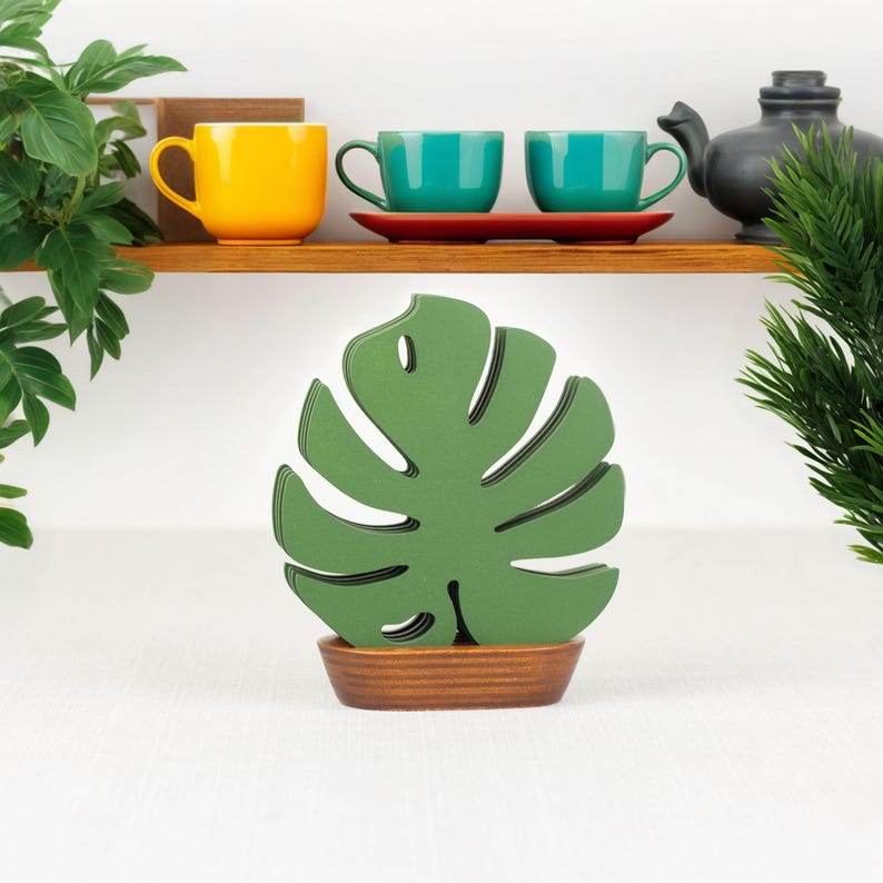 May include: Set of six green monstera leaf-shaped coasters with a brown wooden base. The coasters are stacked on top of each other to form a plant-like shape.