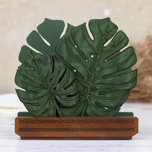 May include: Green wooden napkin holder with a tropical leaf design. The holder has a brown base with a geometric pattern.