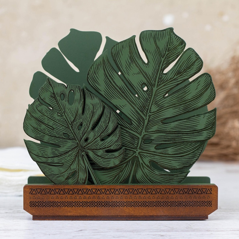 May include: Green wooden napkin holder with a tropical leaf design. The holder has a brown base with a geometric pattern.