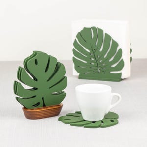 May include: Green monstera leaf shaped coasters and napkin holder. The coasters and napkin holder are made of wood and have a natural wood finish.