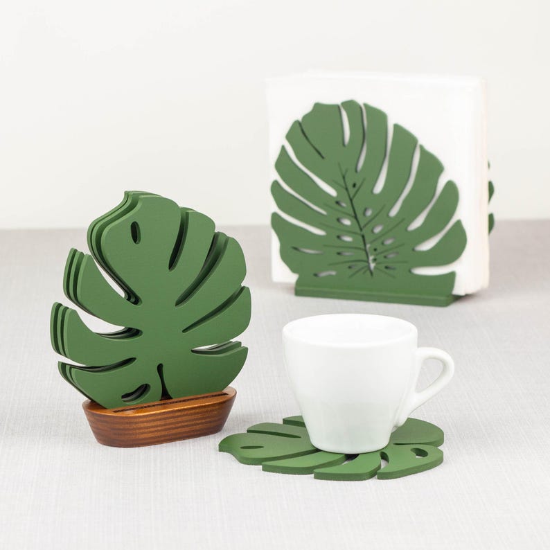 May include: Green monstera leaf shaped coasters and napkin holder. The coasters and napkin holder are made of wood and have a natural wood finish.