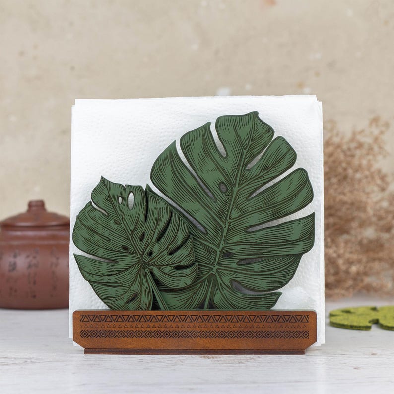 May include: Wooden napkin holder with a green monstera leaf design. The holder has a brown wood base and a decorative border.