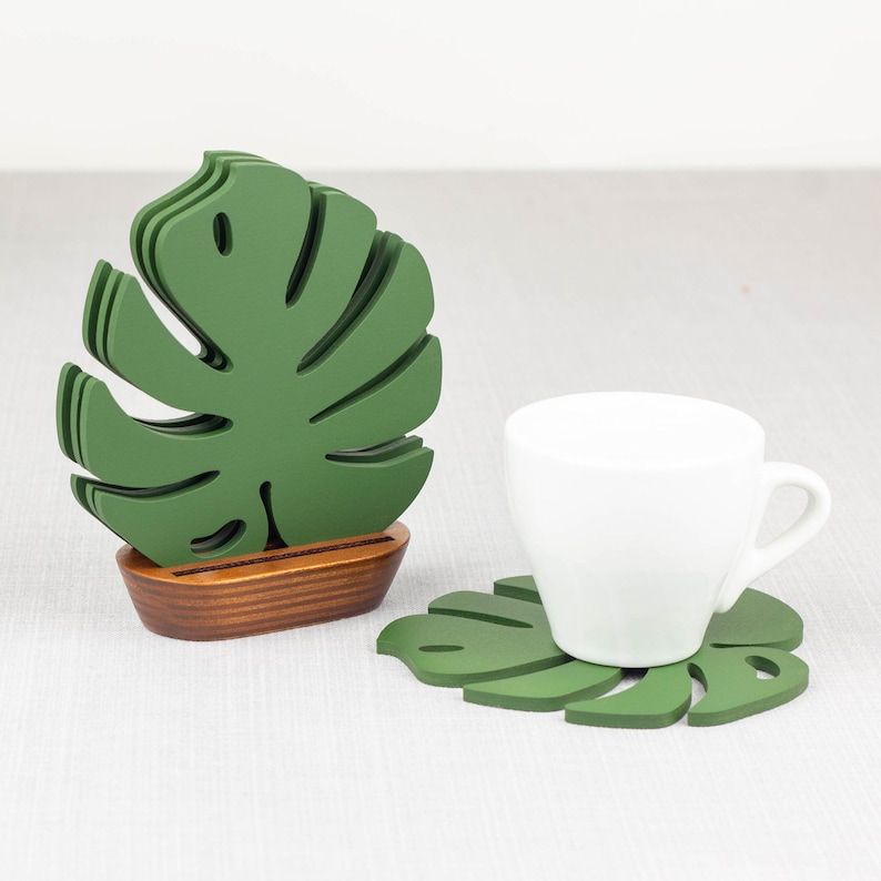 May include: Set of green monstera leaf-shaped coasters. One coaster is stacked on top of the others, and a white coffee cup is resting on a single coaster.