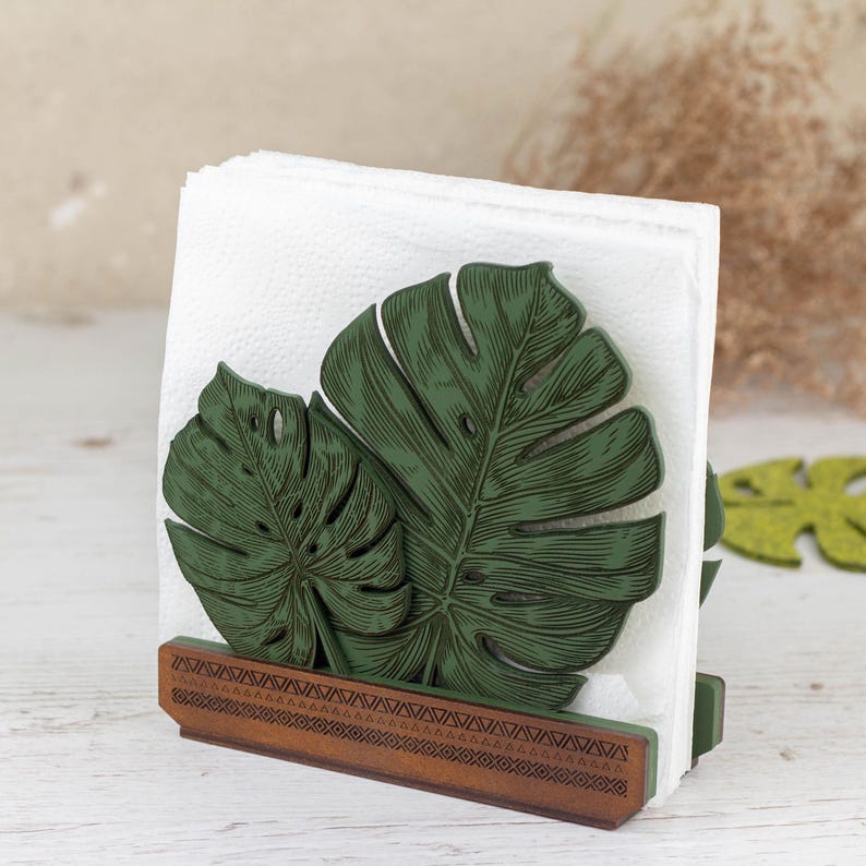 May include: A wooden napkin holder with a green monstera leaf design. The holder has a brown base with a geometric pattern.