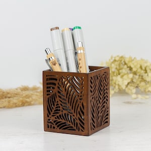 May include: A brown wooden pencil holder with a leaf design. The holder is filled with pens and pencils.