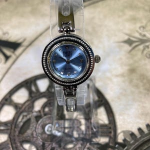 May include: A silver-toned wristwatch with a light blue face and the text "Faded Glory: Quartz." The watch has a round face with a crystal-studded bezel and a silver band. The watch is displayed on a clear stand.