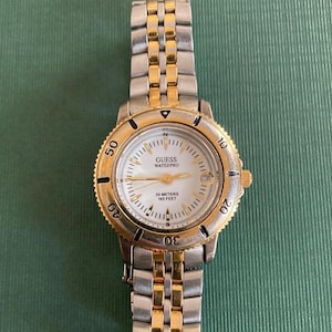 May include: A Guess Waterpro watch with a silver and gold-tone metal band and bezel. The watch face is white with black numerals and hands. The watch is water-resistant to 165 feet.