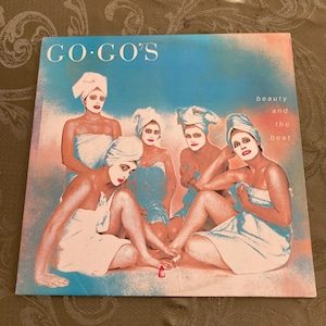 May include: Album cover for the Go-Go's "Beauty and the Beat." The cover features five women in towels and head wraps, with makeup. The background is a mix of blue and pink hues, with the album title and text "beauty and the beat."