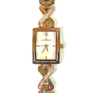 May include: Gold-toned Anne Klein wristwatch with a rectangular face and a cream-colored dial. The watch features Roman numeral XII and the Anne Klein logo. The band has a unique design with diamond accents.