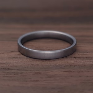 May include: A simple, solid band ring with a smooth, matte finish in a dark charcoal gray color. The ring features a flat, uniform profile with no additional embellishments, engravings, or stones, presenting a clean and understated circular design.