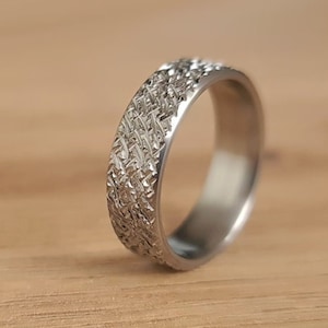 Puede incluir: Un anillo de metal plateado que presenta un patrón texturizado de líneas entrecruzadas en la superficie exterior. La banda tiene un acabado interior liso y pulido, con un ancho constante en toda su circunferencia y un brillo metálico definido.