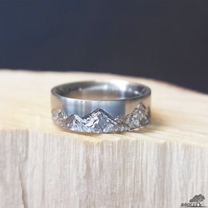May include: A silver-toned metal ring featuring a textured, jagged mountain range relief pattern encircling the outer band. The surface of the ring has a polished finish on the upper portion, while the lower section displays a detailed, rugged landscape design.