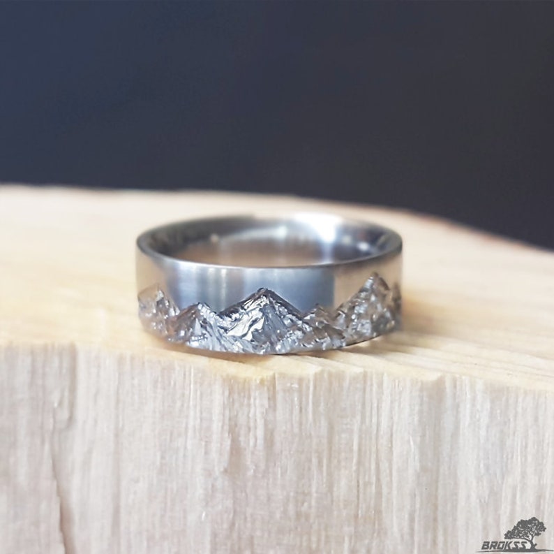 May include: A silver-toned metal ring featuring a textured, jagged mountain range relief pattern encircling the outer band. The surface of the ring has a polished finish on the upper portion, while the lower section displays a detailed, rugged landscape design.