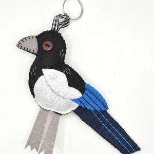 May include: A felt bird keyring featuring a black head and body, a white chest patch, and a long tail with blue accents. The bird has a grey beak, a brown button eye, and two grey felt legs. A black and white gingham ribbon attaches the bird to a metal ring.