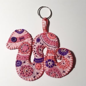 Può includere: Portachiavi a forma di serpente in feltro rosa e viola con un occhio a bottone. Il serpente è arrotolato e ha un nastro gingham rosa e bianco attaccato a un portachiavi argentato.