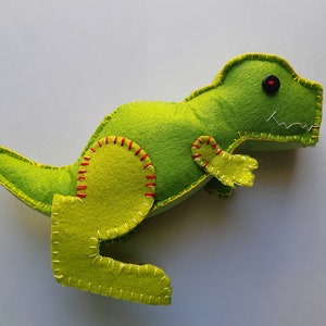 Peut inclure: Jouet dinosaure en feutre vert avec des coutures rouges et jaunes. Le dinosaure a une grosse tête avec un œil en bouton noir et une petite bouche.