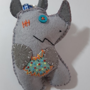 Può includere: Un portachiavi a forma di rinoceronte in feltro grigio con un occhio a bottone blu, cuciture arancioni e una toppa ricamata colorata sul petto.