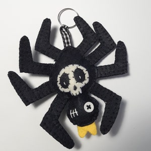 Könnte beinhalten: Schwarzer Filz-Spinnen-Schlüsselanhänger mit einem weißen Totenkopf- und Knochen-Design. Die Spinne hat eine gelbe Krone und ein Knopfauge.
