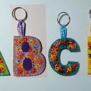 May include: Four felt letter keychains with embroidered flowers. The letters are A, B, C, and D. The keychains are in green, purple, blue, and yellow.