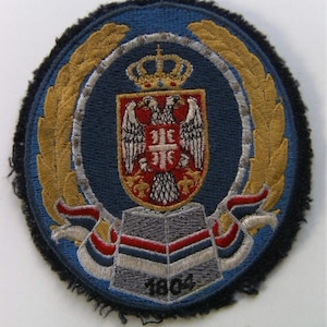 Pode incluir: Um remendo bordado azul e dourado com um brasão de armas que apresenta uma coroa, duas águilas e um escudo com uma cruz. O escudo é rodeado por uma coroa de louro. O ano 1804 está impresso abaixo do escudo.