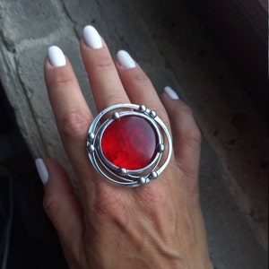 May include: A silver ring featuring a large, round, red glass stone set within a circular frame. The frame has a series of concentric circles and small silver beads.