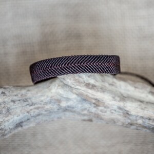 Könnte beinhalten: Ein dunkelbraunes gewebtes Armband mit Fischgrätenmuster. Das Armband liegt auf einem Stück Treibholz.
