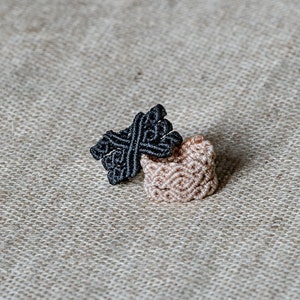 Op de afbeelding: Twee macramé ringen, één zwart en één beige, op een textuurrijke beige stoffen achtergrond.