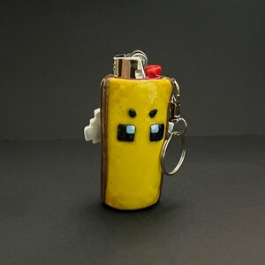 May include: A yellow lighter case with a brown border and a character face featuring black square eyes with blue accents and small black eyebrows. The case includes a silver metal keychain attachment and a red ignition button on the top.