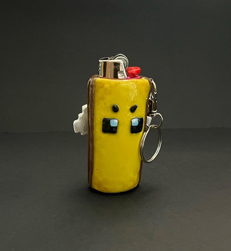 May include: A yellow lighter case with a brown border and a character face featuring black square eyes with blue accents and small black eyebrows. The case includes a silver metal keychain attachment and a red ignition button on the top.