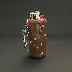 May include: A brown lighter case with a smooth finish, decorated with small, colorful, spherical accents in red, blue, green, orange, and yellow. The lighter features a silver metal top and a red ignition button, with a silver metal keychain ring attached.