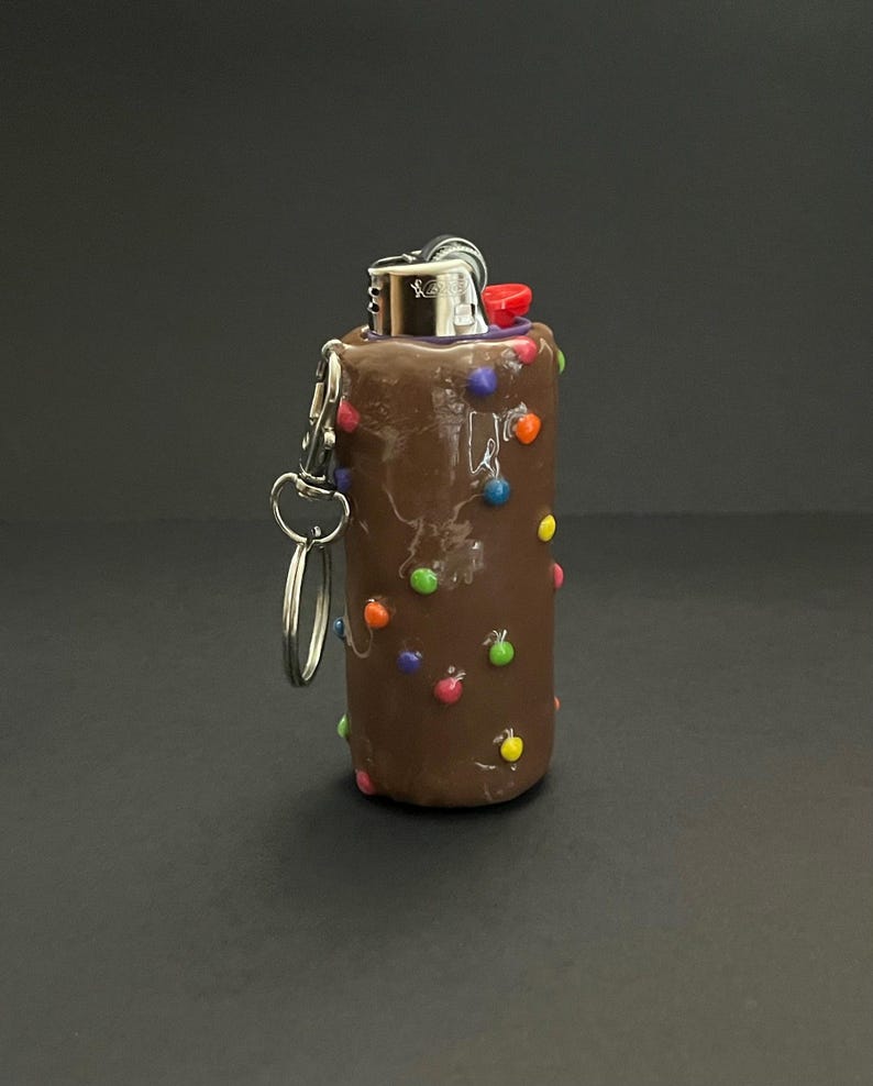 May include: A brown lighter case with a smooth finish, decorated with small, colorful, spherical accents in red, blue, green, orange, and yellow. The lighter features a silver metal top and a red ignition button, with a silver metal keychain ring attached.