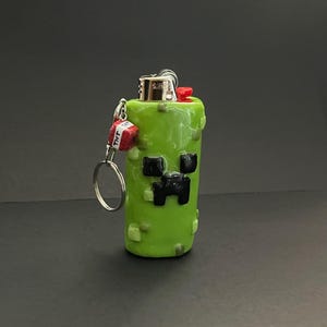 May include: A lime green lighter case featuring a pixelated black face design, textured with small green squares. A silver metal keychain is attached to the side, holding a small red cube charm with the white text TNT printed on its sides.