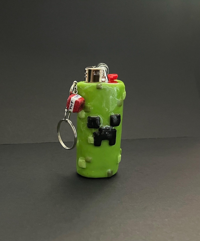 May include: A lime green lighter case featuring a pixelated black face design, textured with small green squares. A silver metal keychain is attached to the side, holding a small red cube charm with the white text TNT printed on its sides.